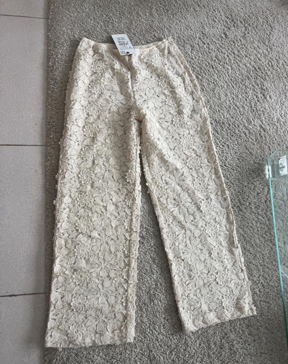 Cream floral lace trousers with tags, laid out on a grey rug next to a tiled floor.