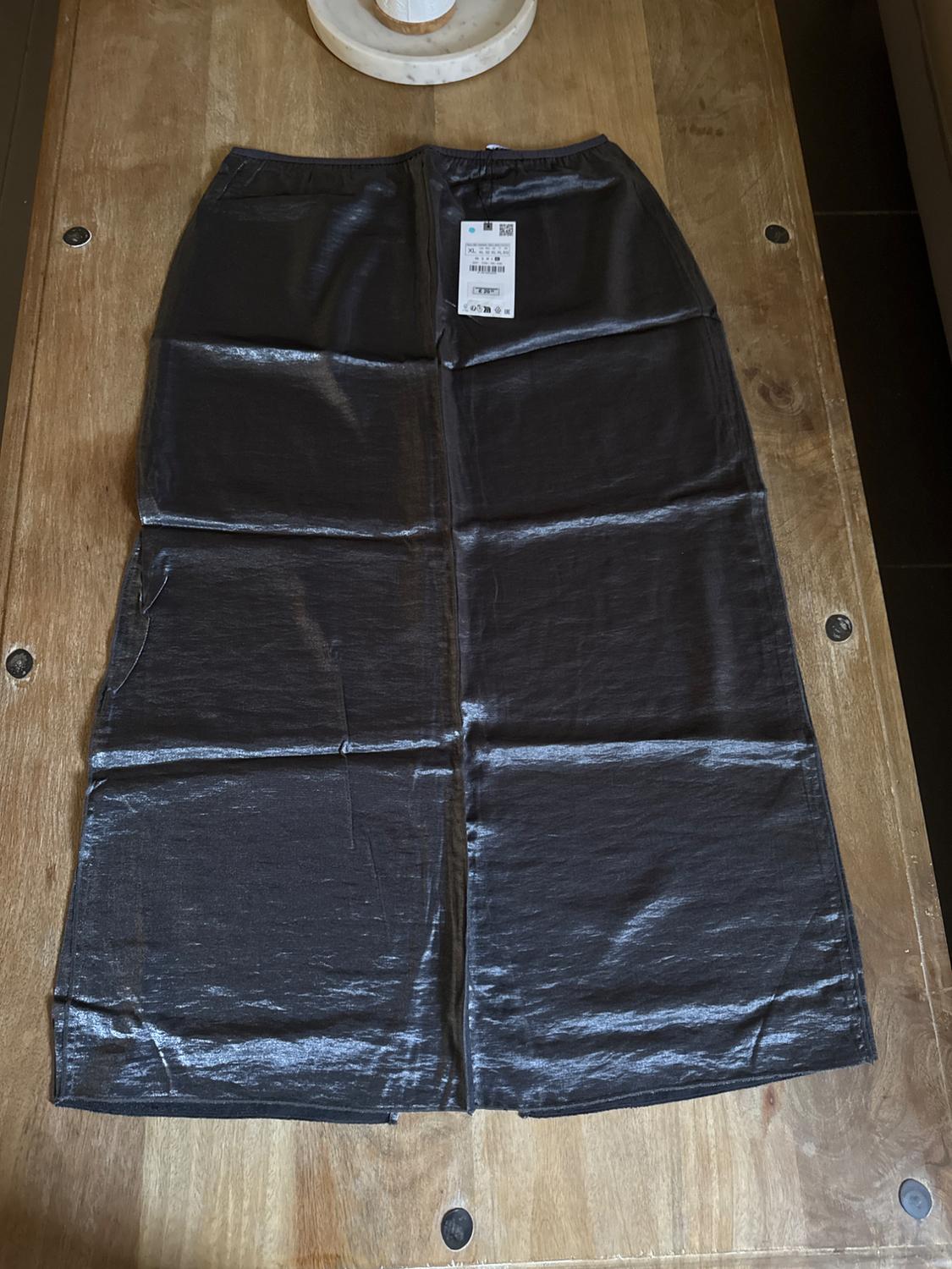 Long black skirt made of shiny fabric on a wooden table. White label visible on the top centre of the skirt.