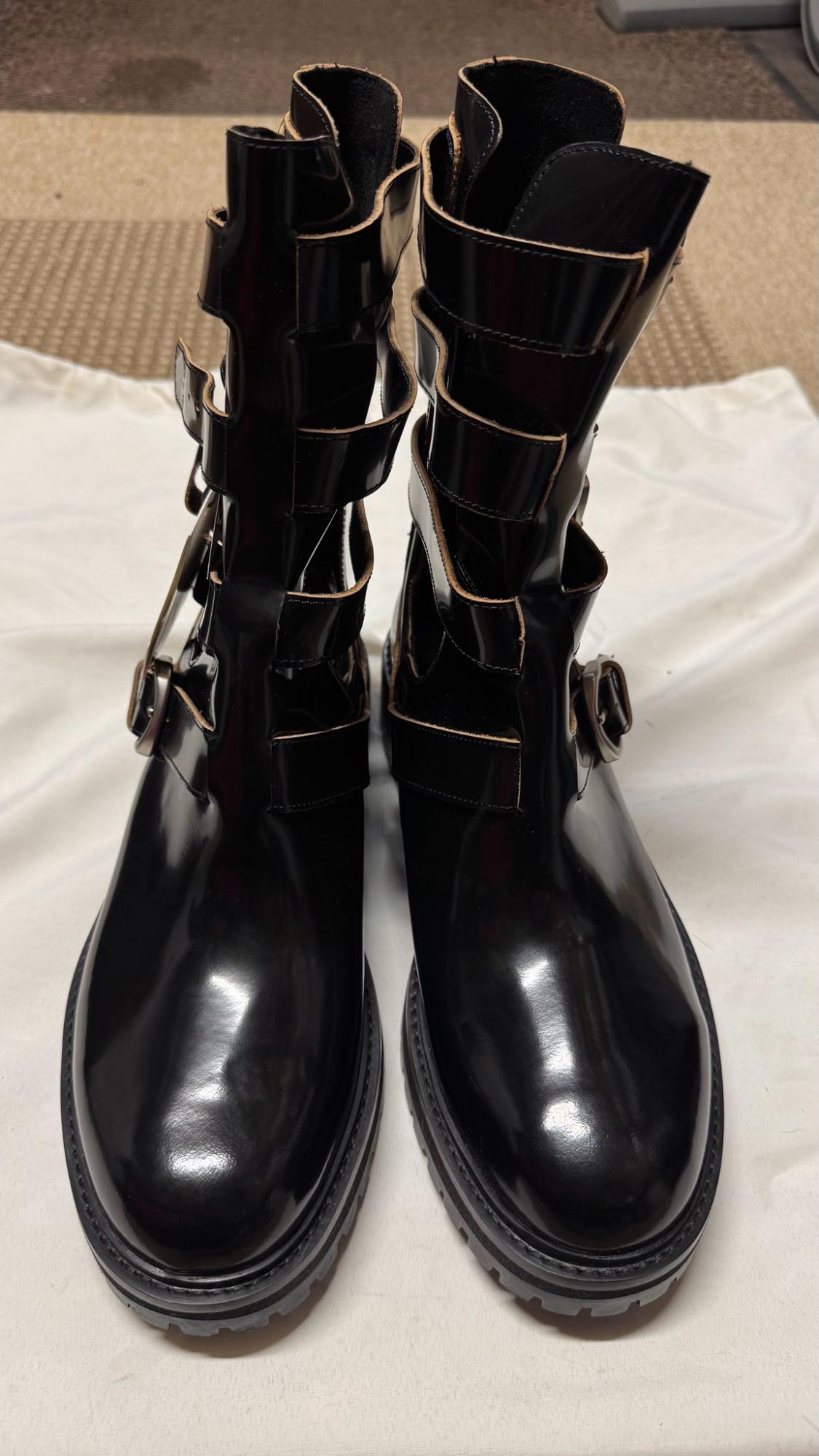 Shiny black boots with multiple straps, side buckles and a thick, chunky sole, resting on a white fabric.