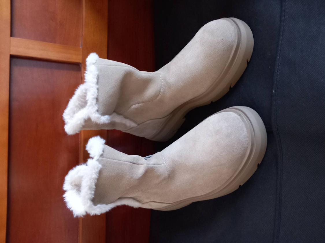 Beige suede boots with white plush lining, on a dark wood and fabric background.