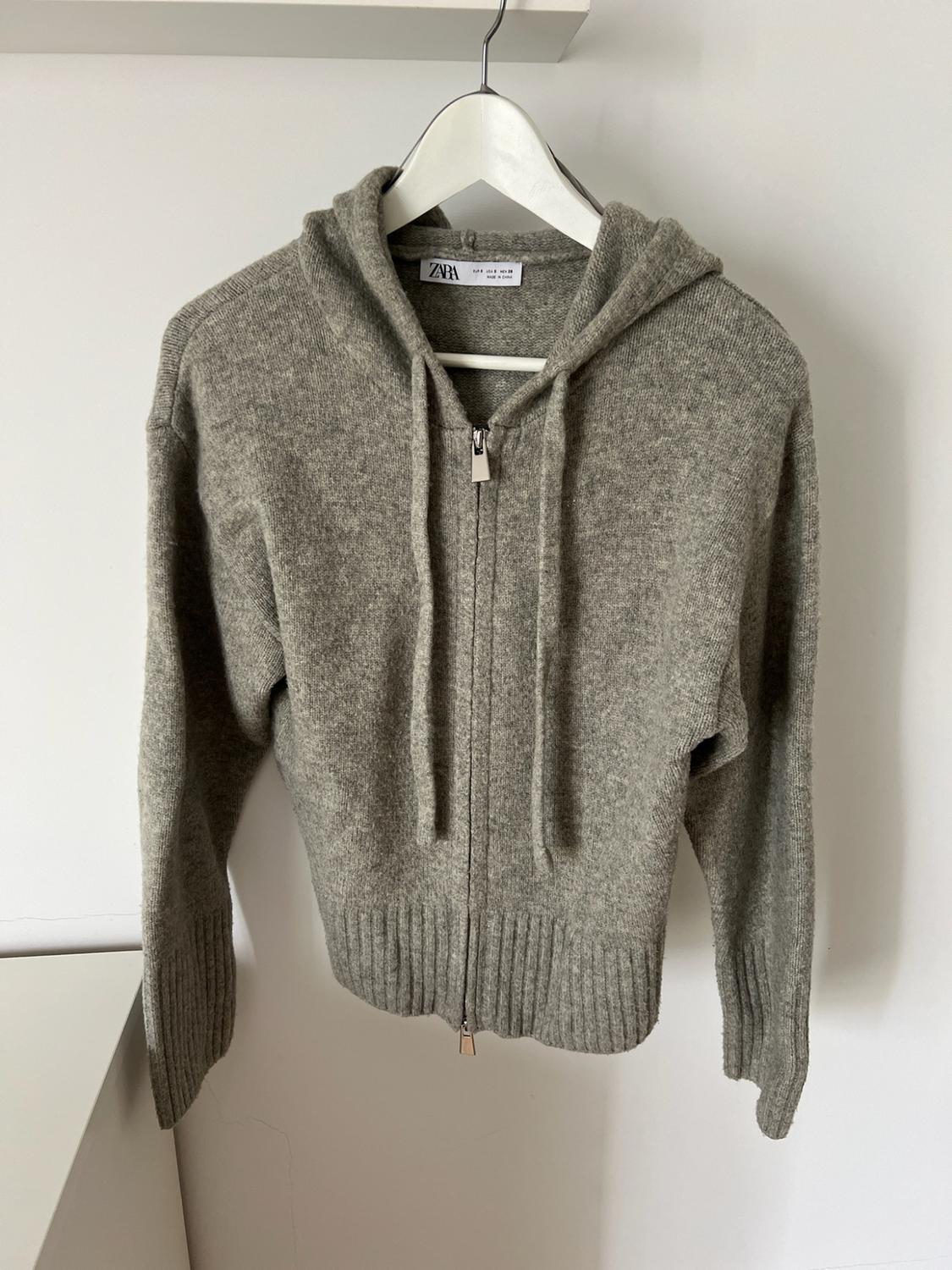 Grey knitted hooded sweatshirt with zip and drawstring, hanging on a white hanger against a white wall. ZARA label.