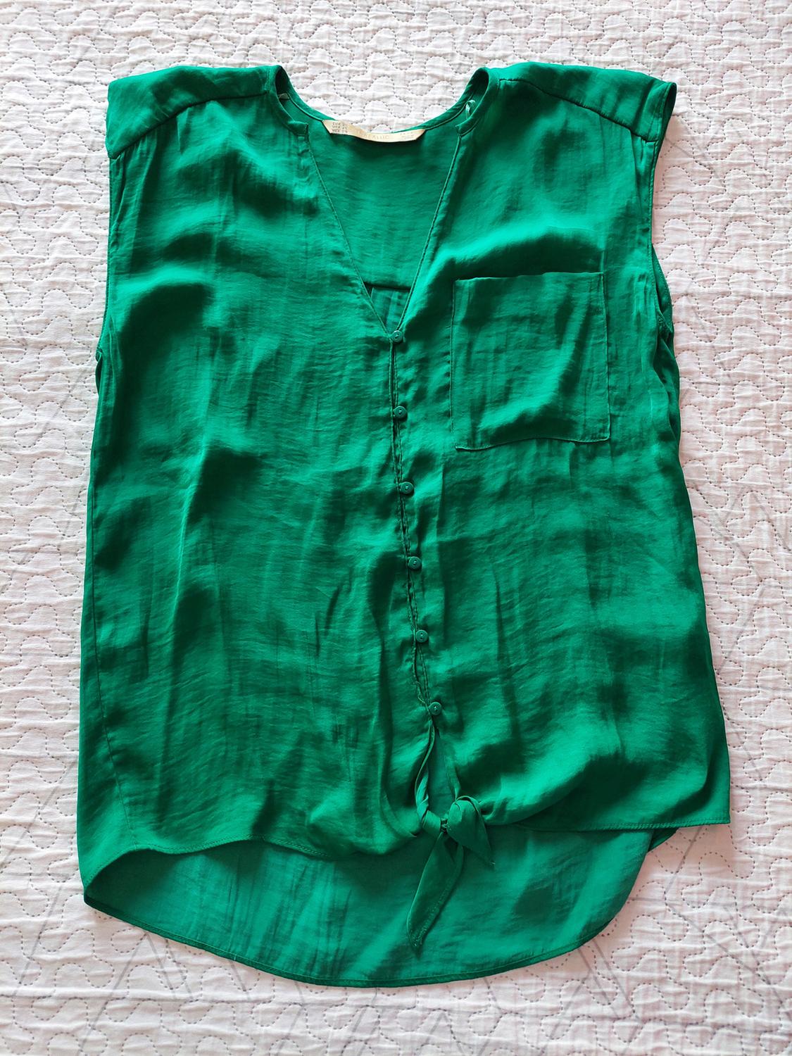 Green sleeveless shirt, V-neck, front buttons, chest pocket and tied hem, on a textured white surface.