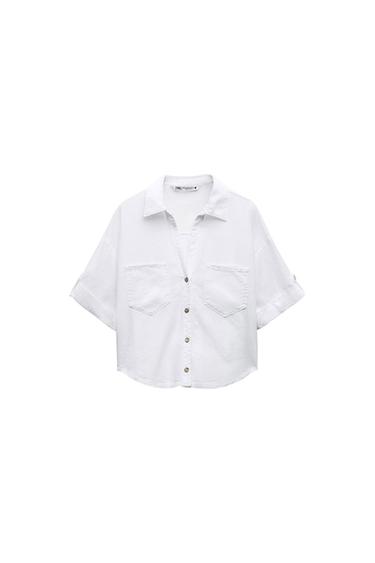 White short-sleeved shirt with brown buttons and two front pockets. Visible label on the collar. White background.
