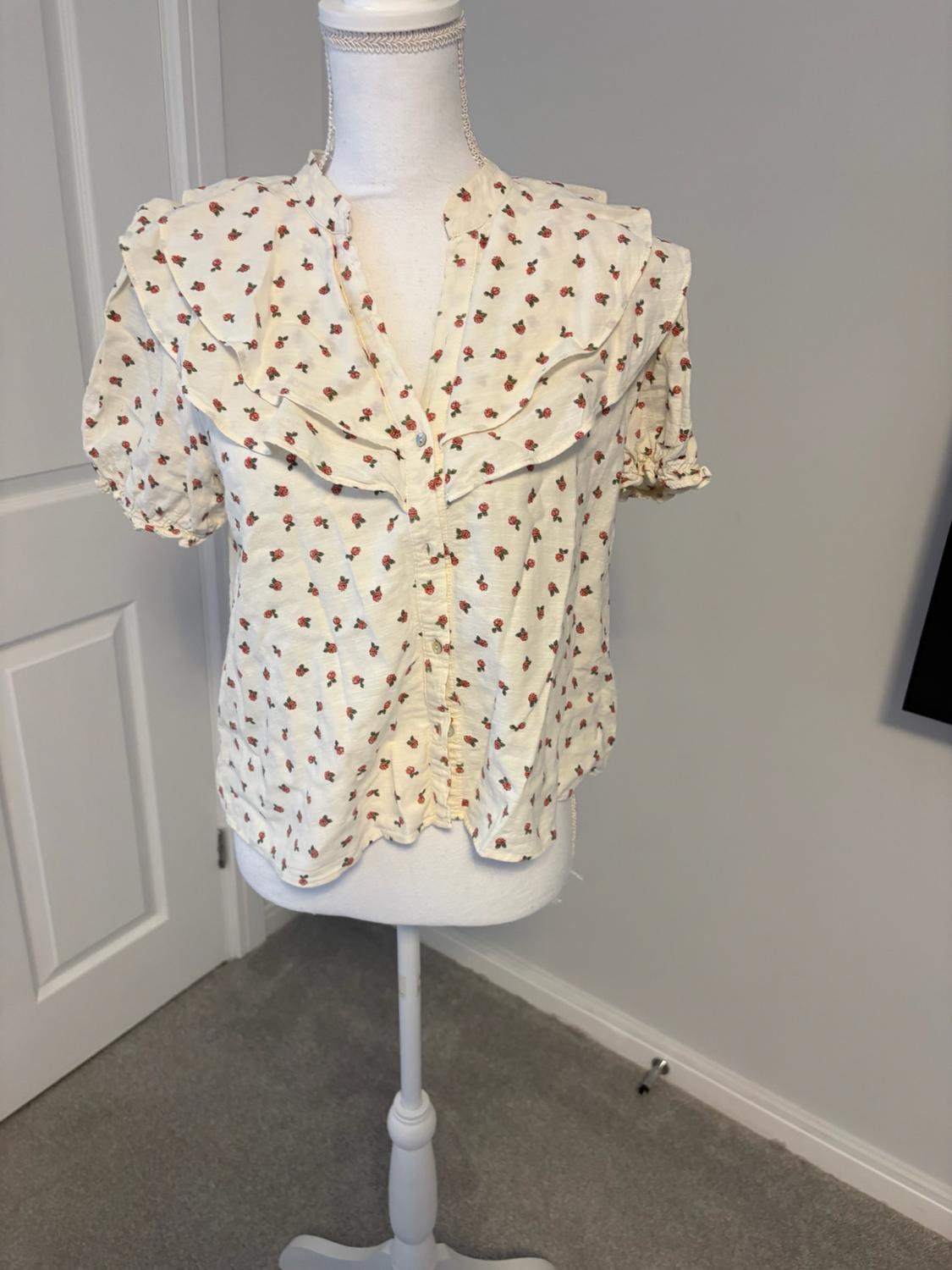 Mannequin indoors wearing a cream blouse with a red floral print, V-neck with frills, front buttons and short puff sleeves.