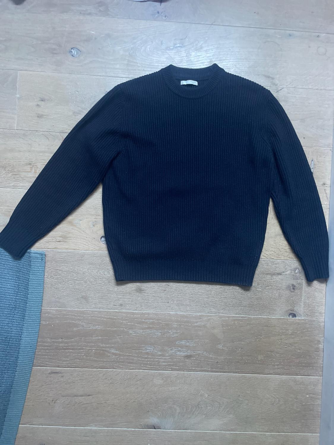 Dark ribbed-knit jumper with long sleeves and a round neck, laid out on a light wooden floor next to a blue rug.