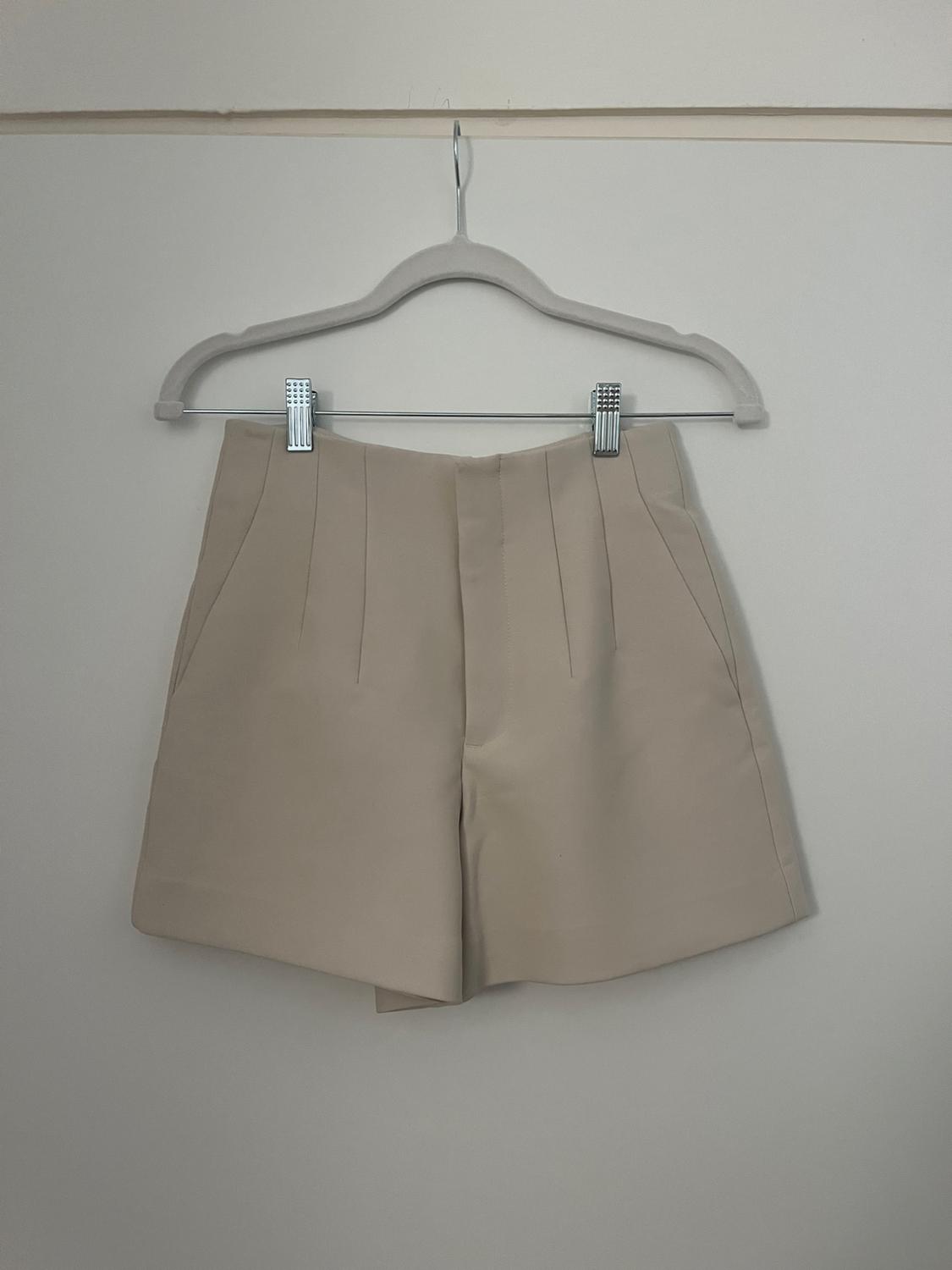 Beige shorts hanging on a grey hanger against a white background. Simple design with front pleats.