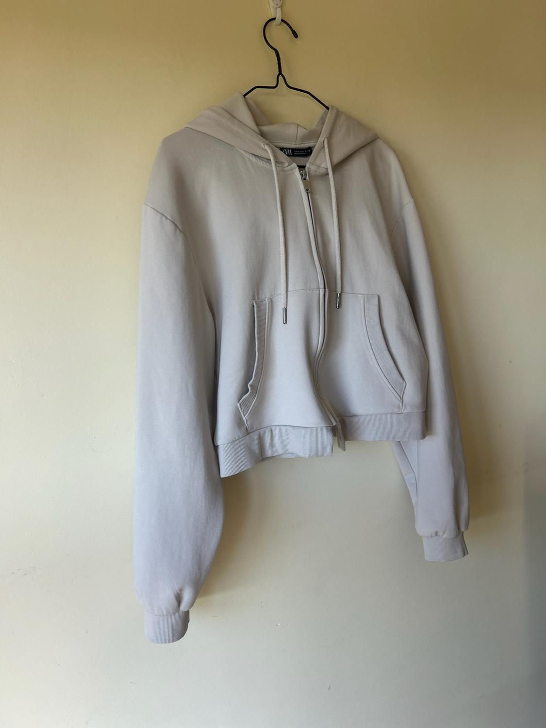 Basic light beige hoodie with a hood, zip and pockets, hanging from a black hanger against a smooth, neutral-toned wall.