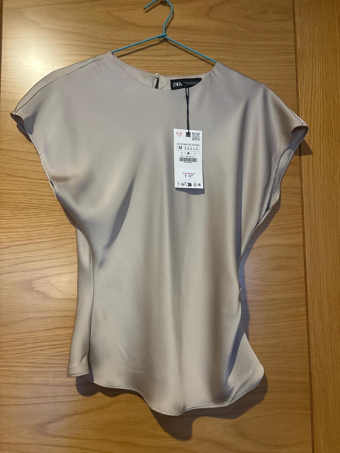 Beige satin blouse with short sleeves hanging on a blue hanger on a wooden door. Label with the text ZARA, M, €22.