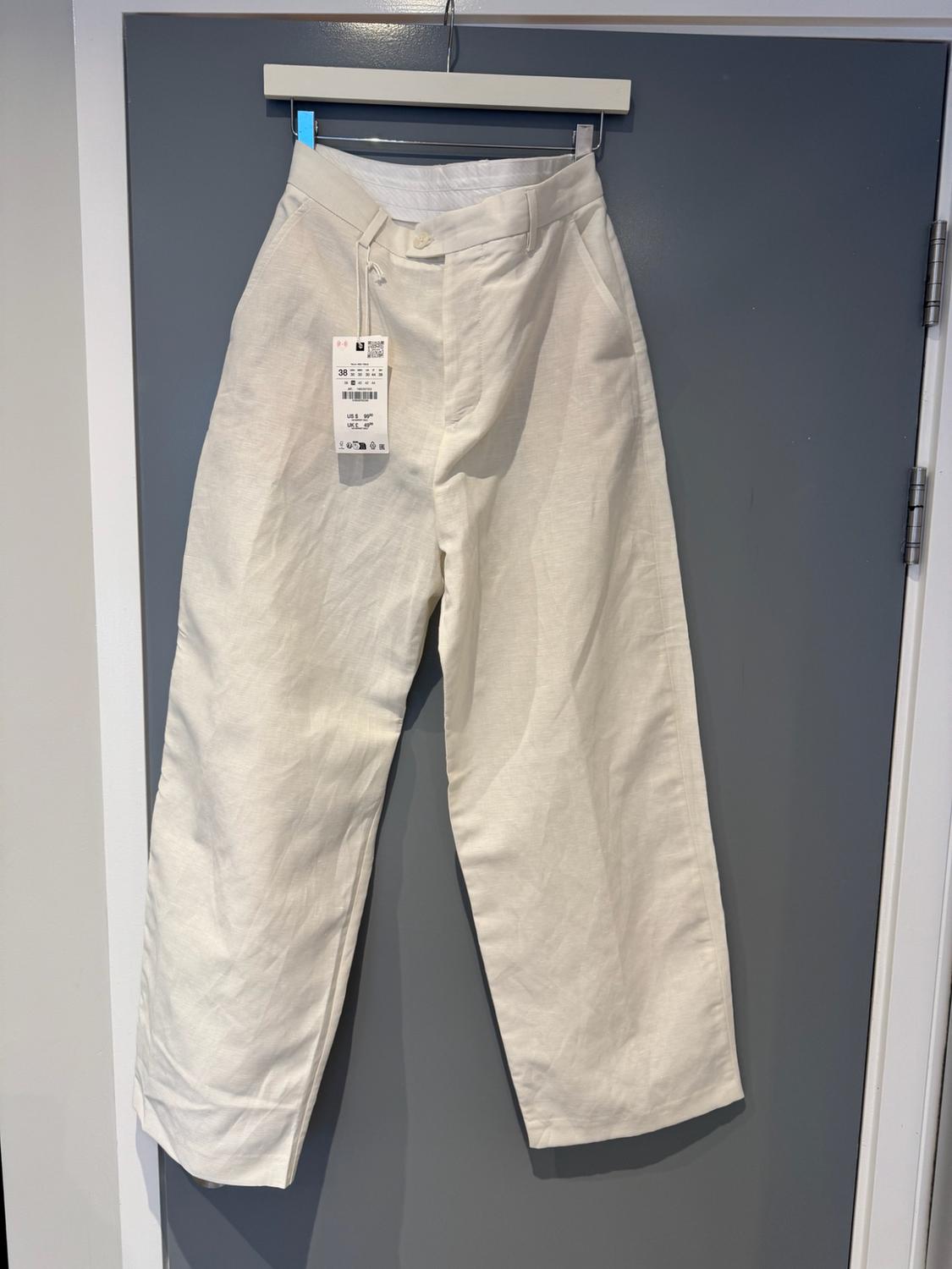 White long trousers hanging on a coat hook over a grey door. There is a visible label at the waist.
