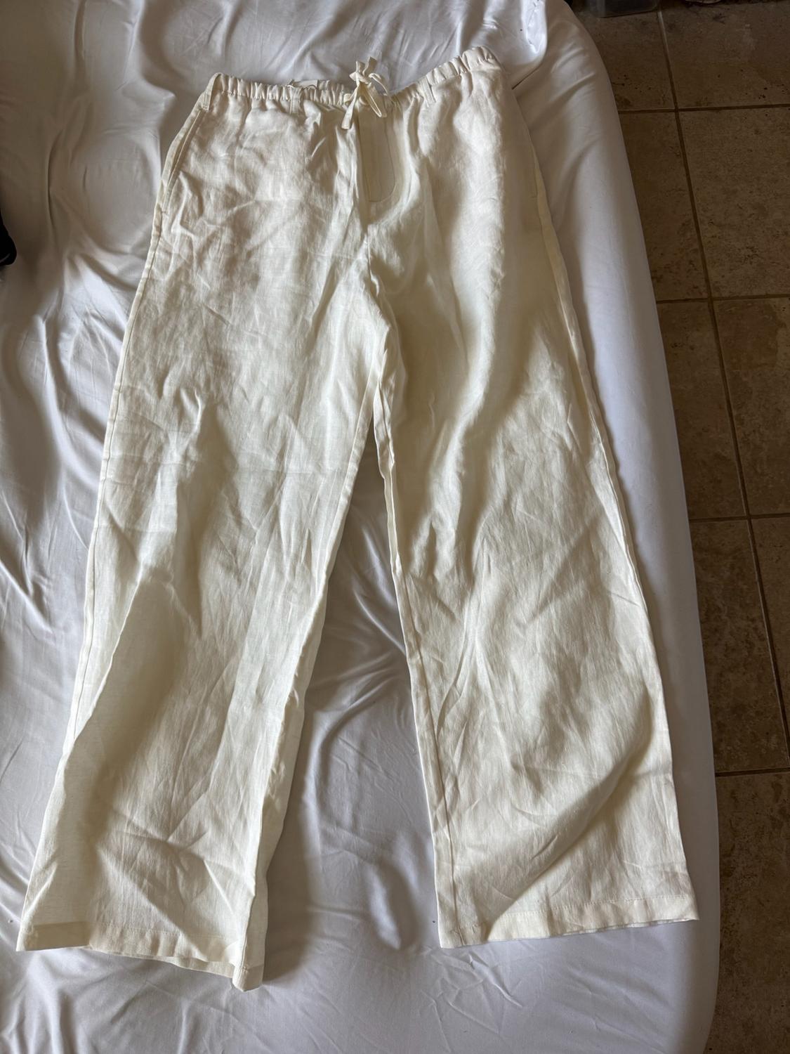 White wide-leg trousers with an elasticated waistband and drawstring, laid out on a bed with white sheets. Casual, relaxed style.