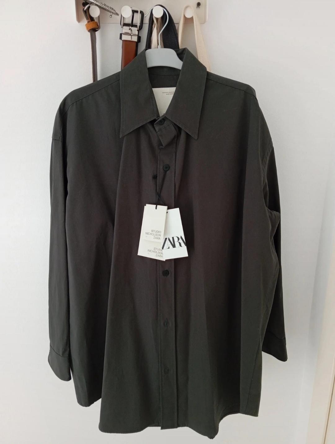 Long-sleeved black shirt hanging on a white hanger, with labels that say "STUDIO NICHOLSON ZARA", background with hanging belts.