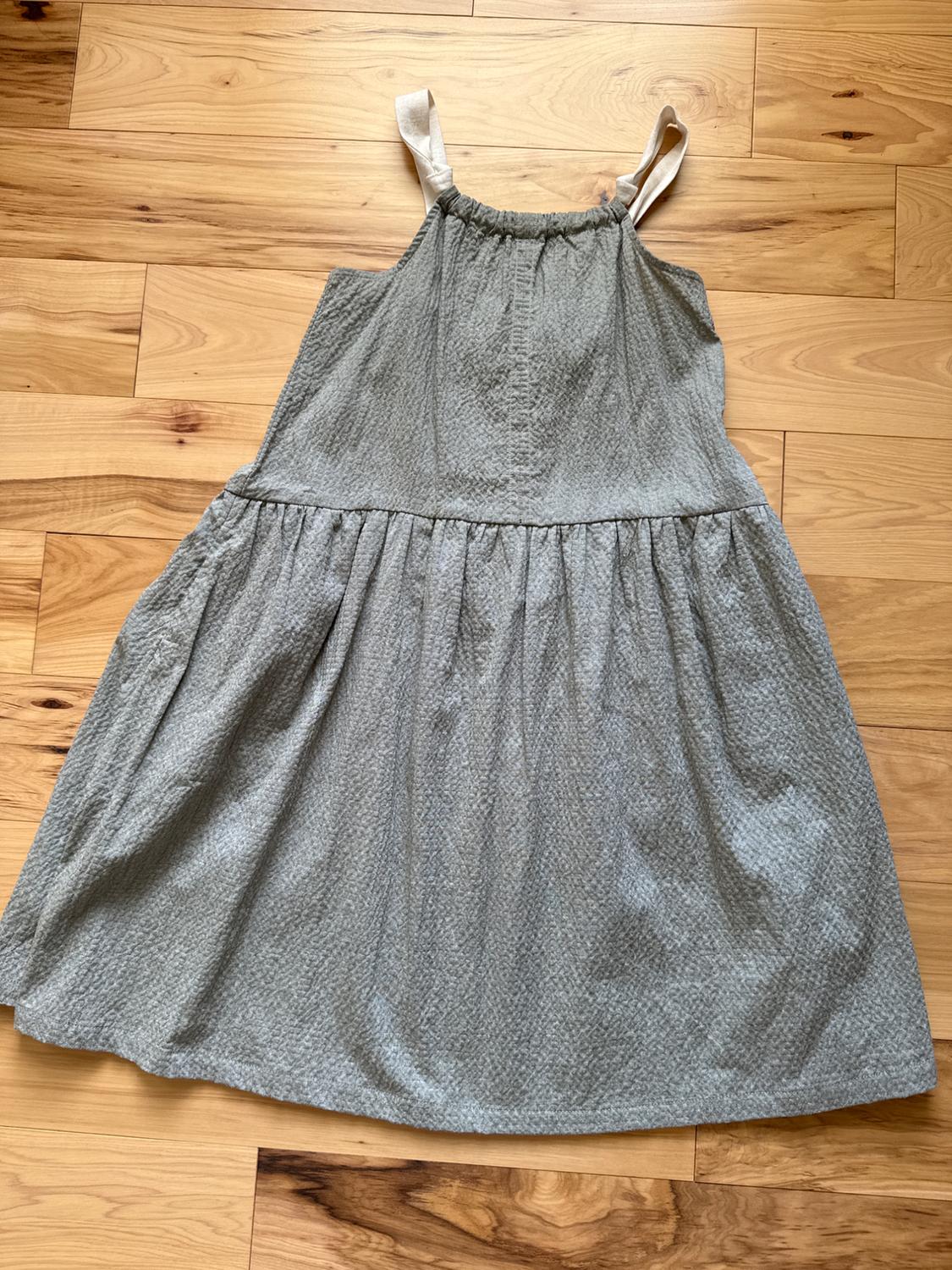 Sleeveless gray dress with a light texture, adjustable beige fabric straps, on a light wood floor with visible grain.
