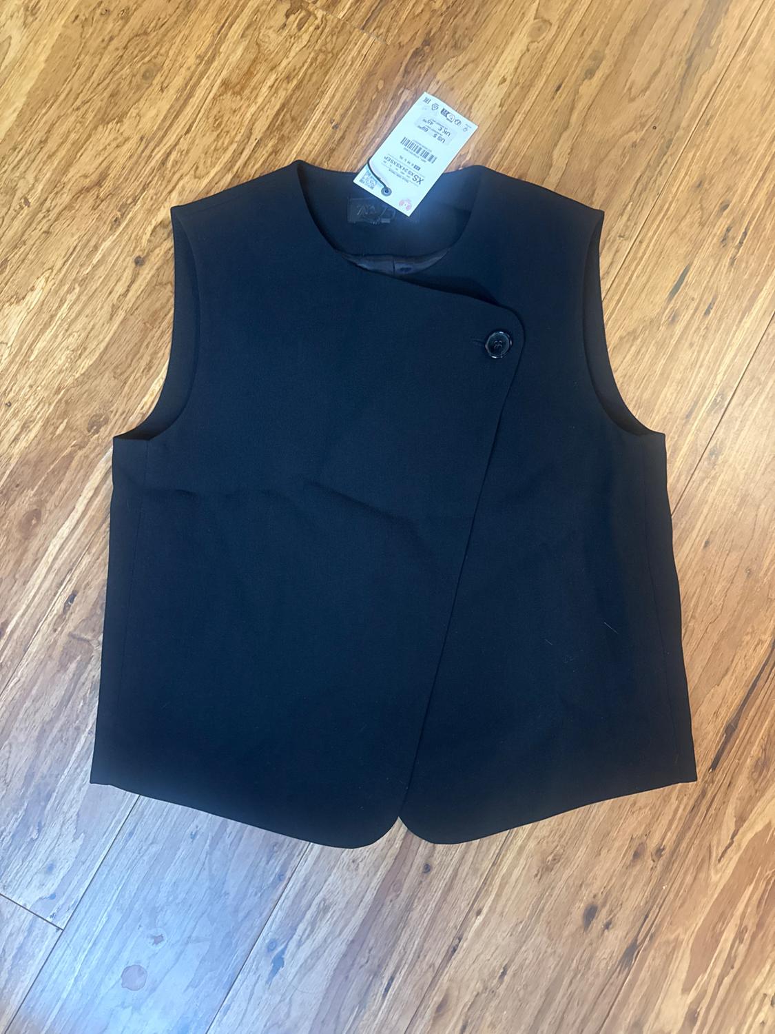 Black sleeveless vest with an asymmetrical zipper and a single button, laid out on a wooden floor.