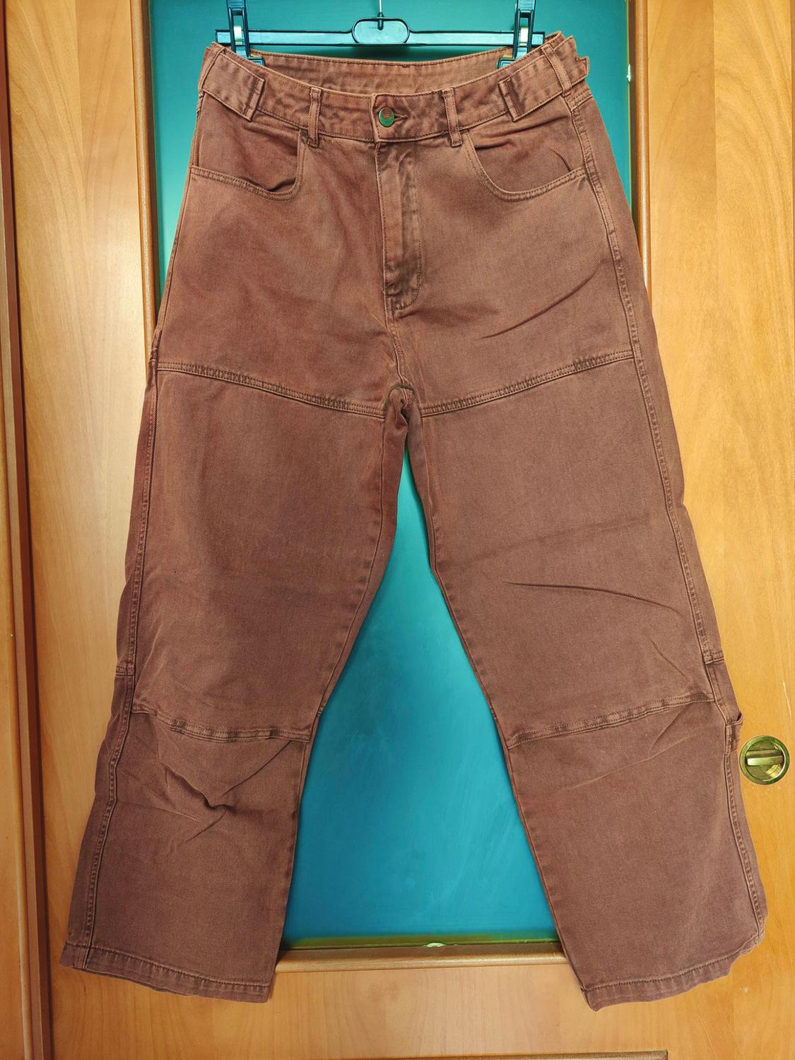 Brown trousers with horizontal stitching hanging from a green coat hanger over a wooden door with blue glass.