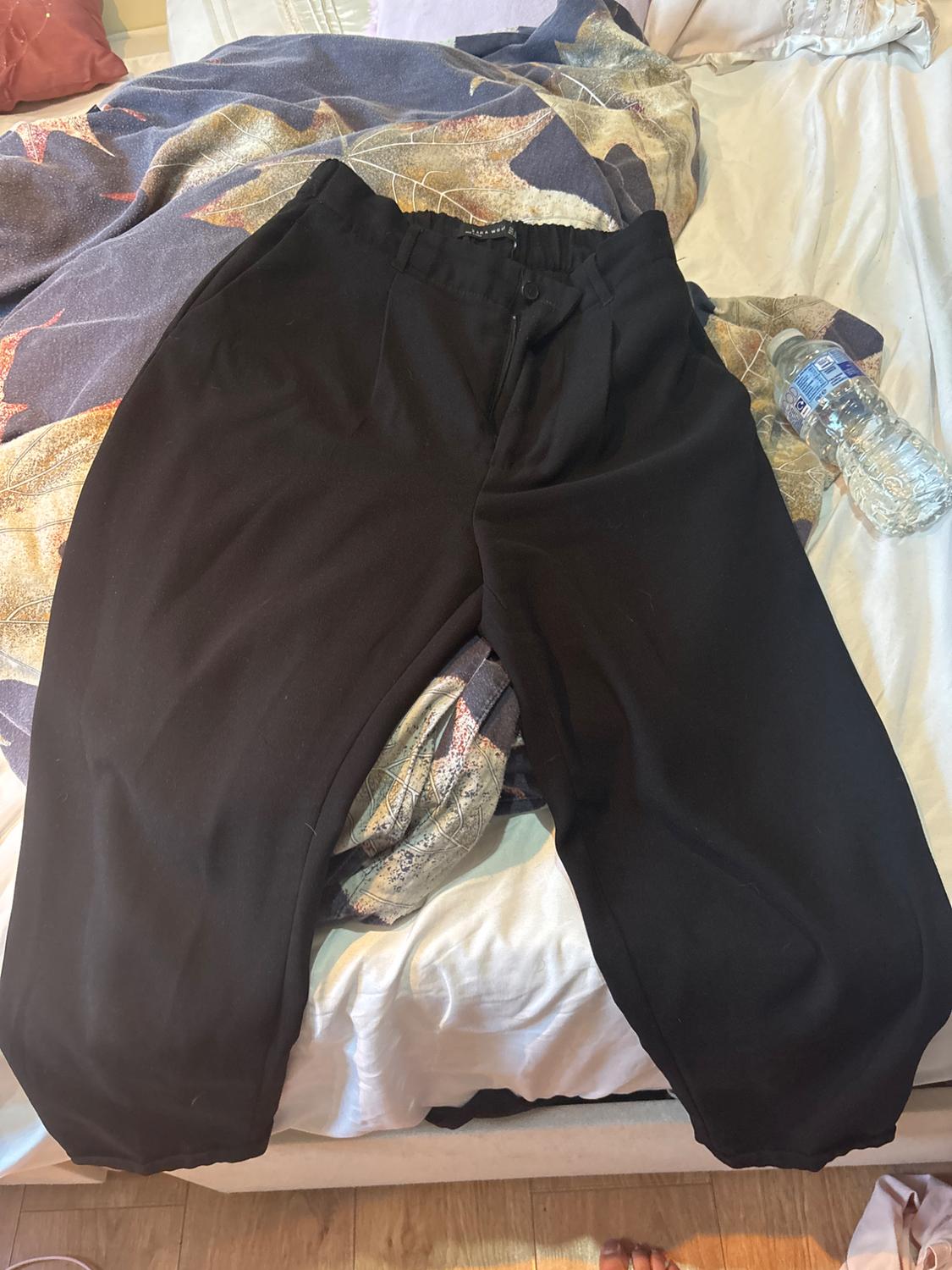 ZARA MAN black trousers on an unmade bed with white sheets and a leaf-print throw. Relaxed atmosphere next to a bottle of water.