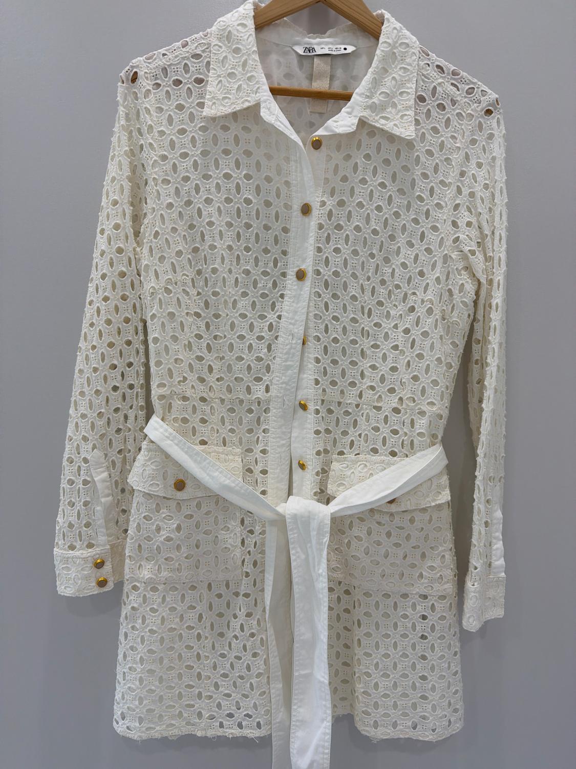 White lace dress with gold buttons and a belt, hanging on a wooden hanger. Visible tag: Zara.