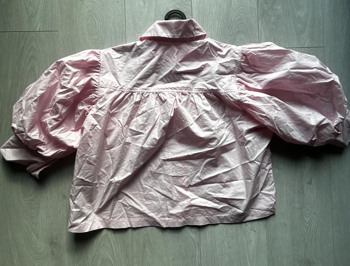 Light pink crinkled blouse with puff sleeves and gathered detail at the back, laid out on a grey wooden floor.