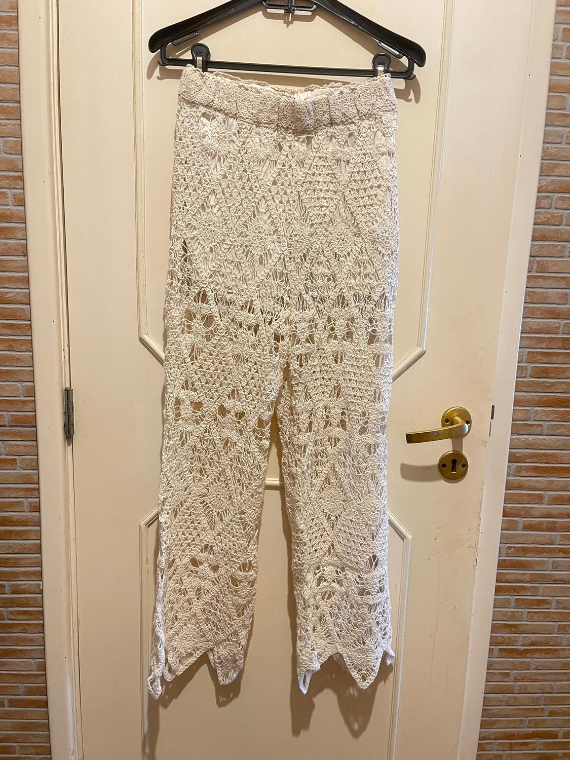 White crochet trousers hanging on a black hanger over a beige door, with a brick wall background.