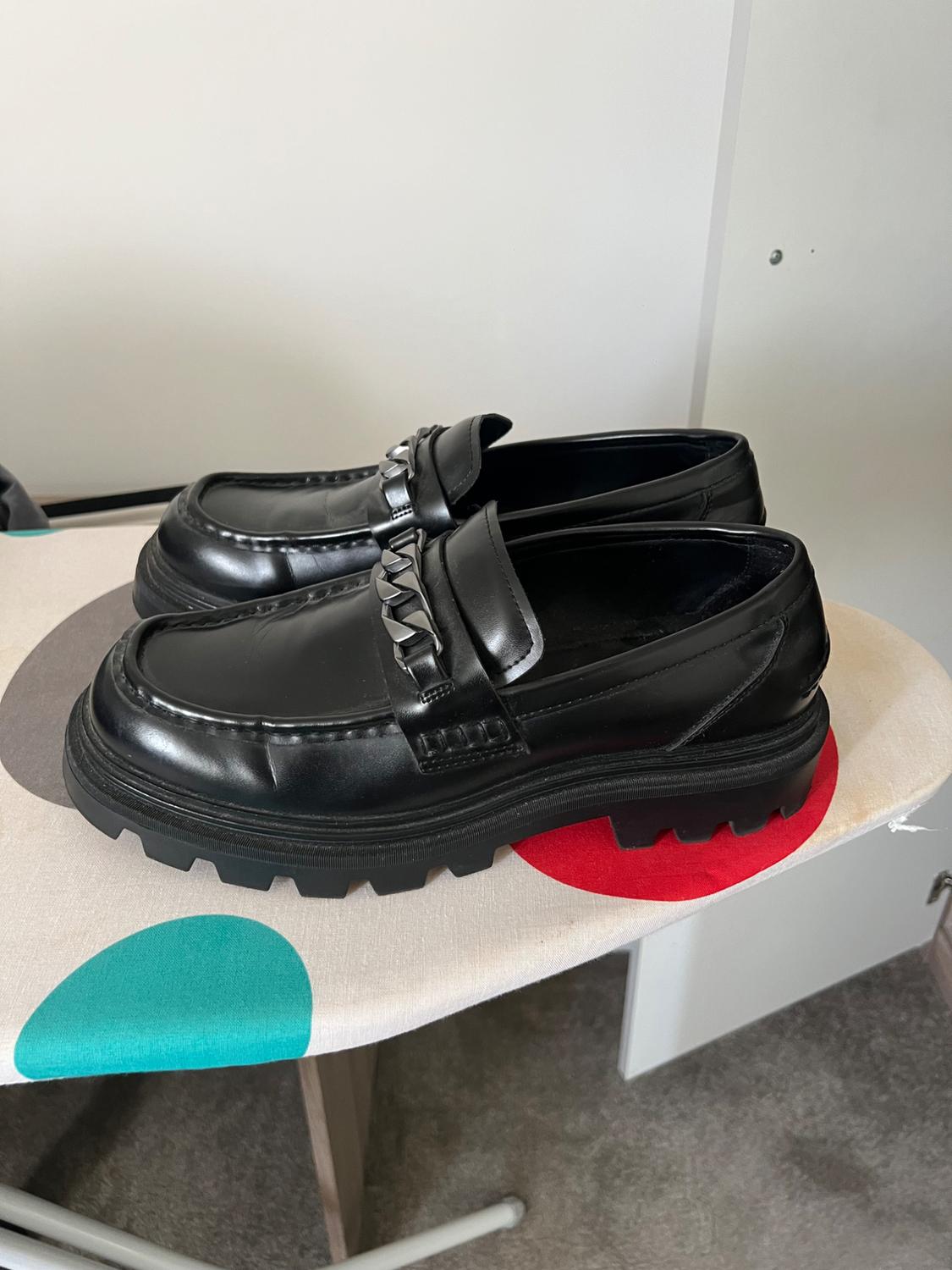 Pair of black chunky-soled loafers with a silver chain, resting on an ironing board with a cover featuring large colourful polka dots.