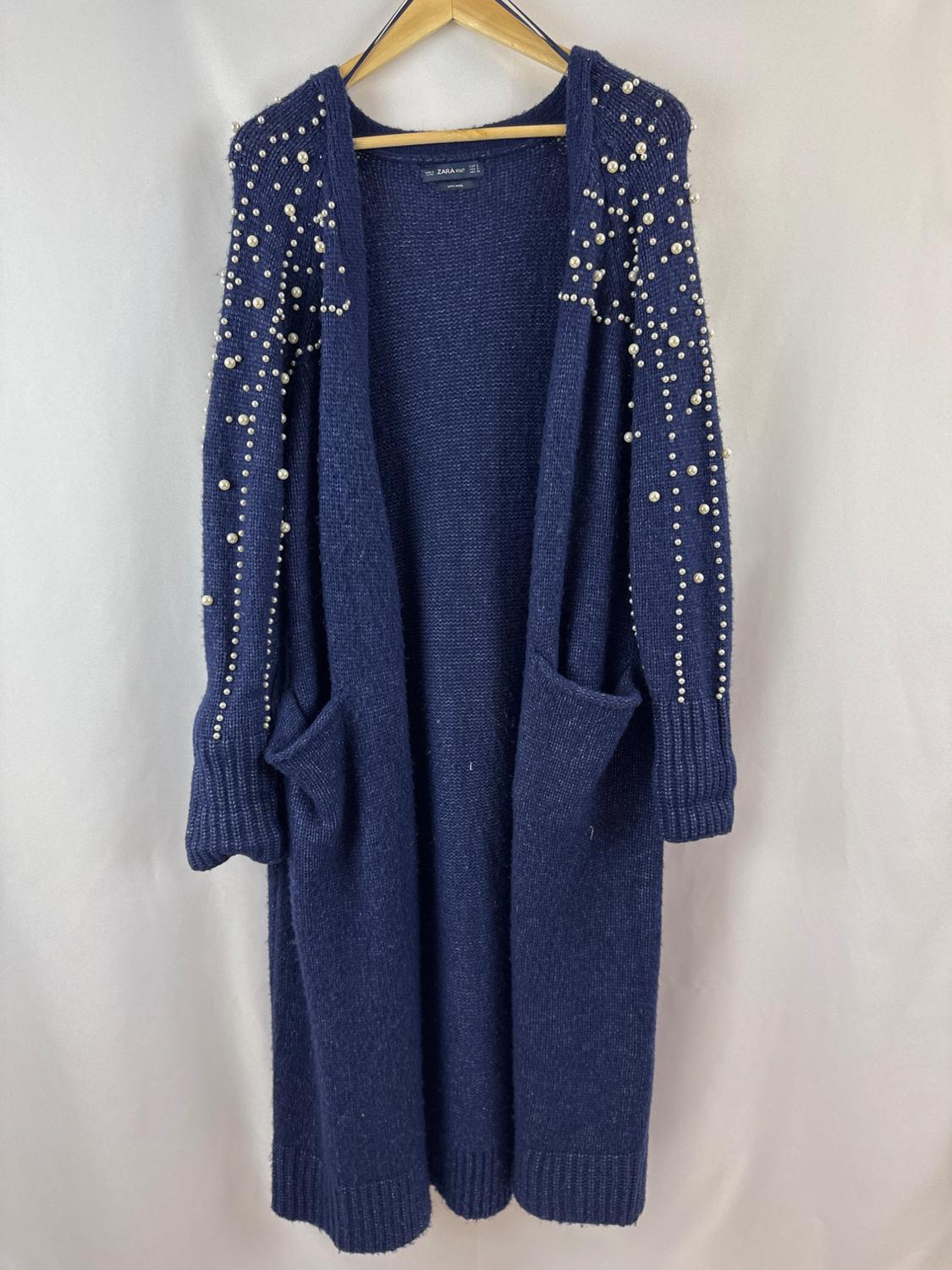 Front view of a blue cardigan with pearls on the sleeves and side pockets.