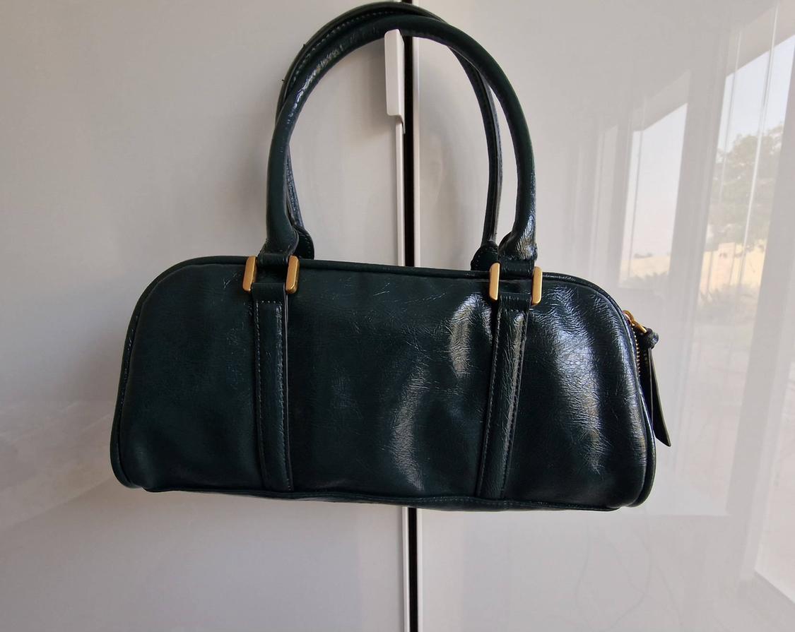 Dark green handbag with a glossy finish, two handles and gold detailing, hanging in a white wardrobe.
