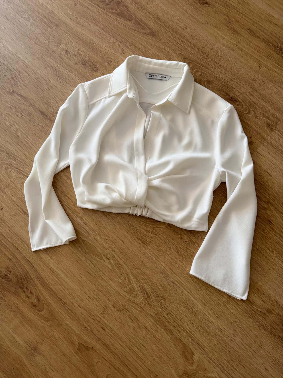 White cropped long-sleeved shirt with a collar and front knot, laid out on a wooden floor. Label with the text ZARA.