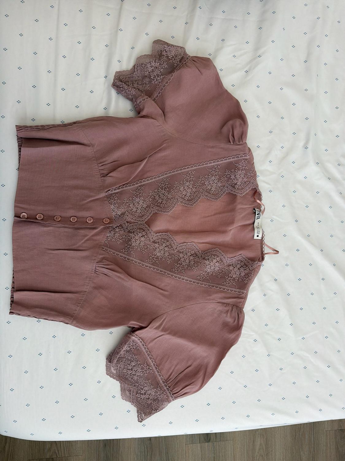 Pink short-sleeved blouse with lace, front buttons and ZARA label, laid out on a white sheet with small blue dots.