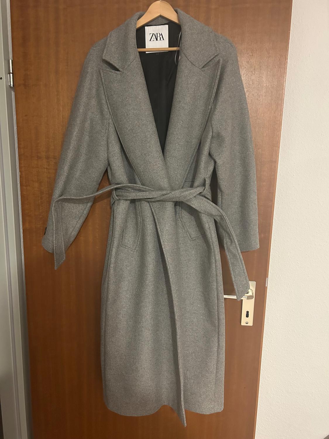 Grey coat with belt hanging on a wooden door. Visible label with the text "ZARA".