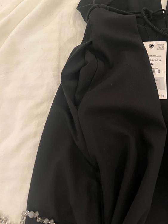 Black fabric on a white surface with a visible label. A transparent beaded necklace can be seen in the bottom left corner.