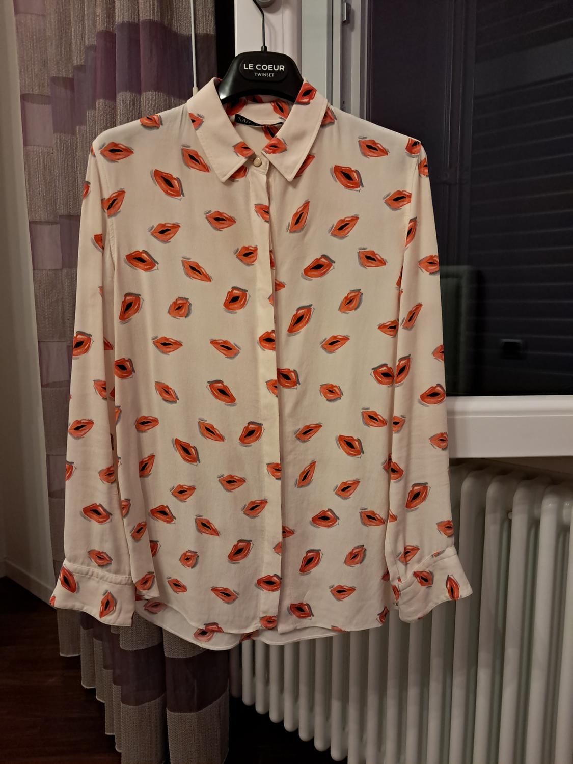 Here is the sentence rephrased while maintaining the same meaning and context, and complying with all the restrictions: A white shirt patterned with orange lips, hanging on a hanger displaying the text LE COEUR TWINSET, positioned in front of a window and a radiator.