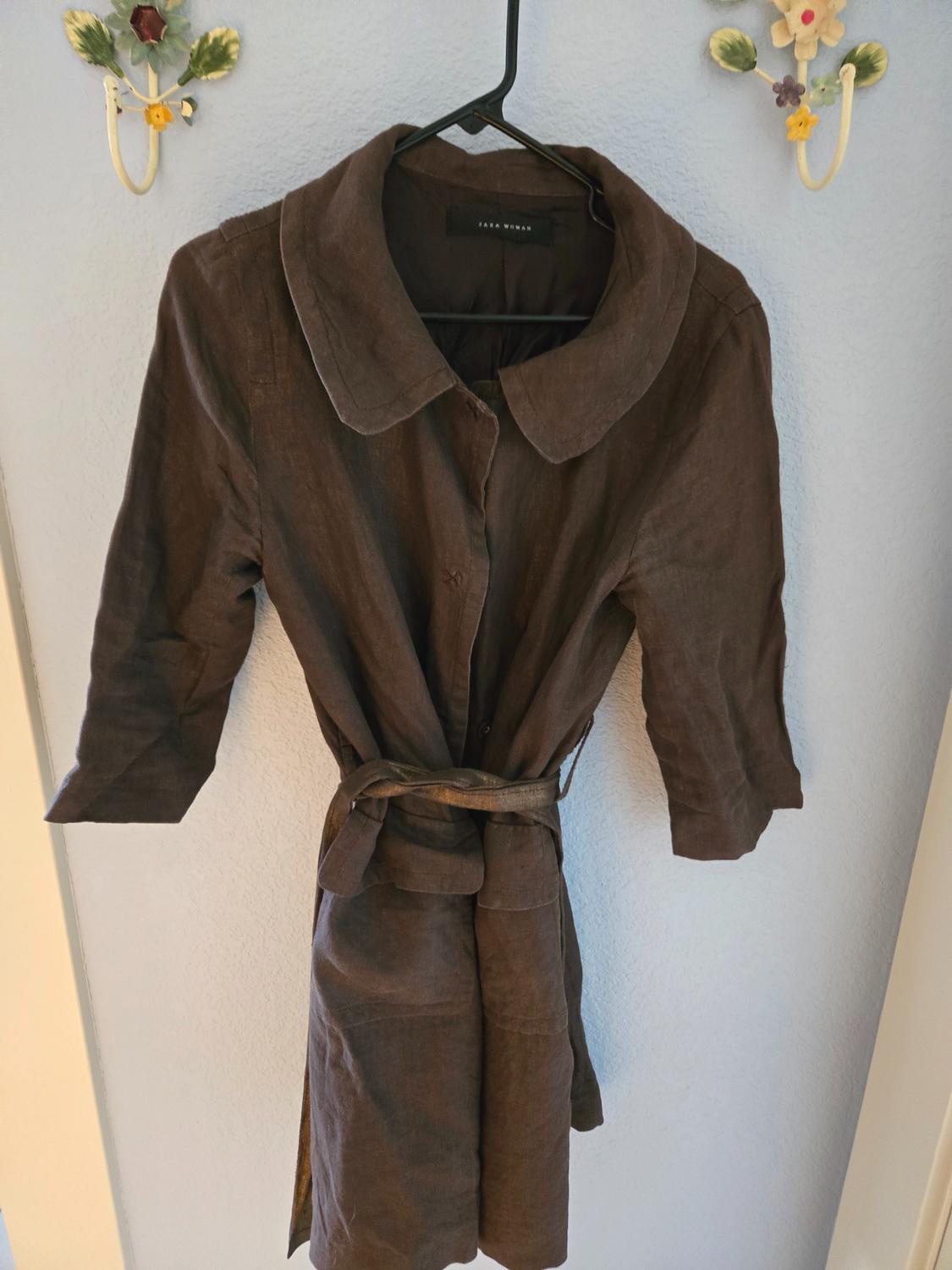 Brown three-quarter sleeve dress hanging on a black hanger, with a matching belt. Background with a light wall and decorative hooks.