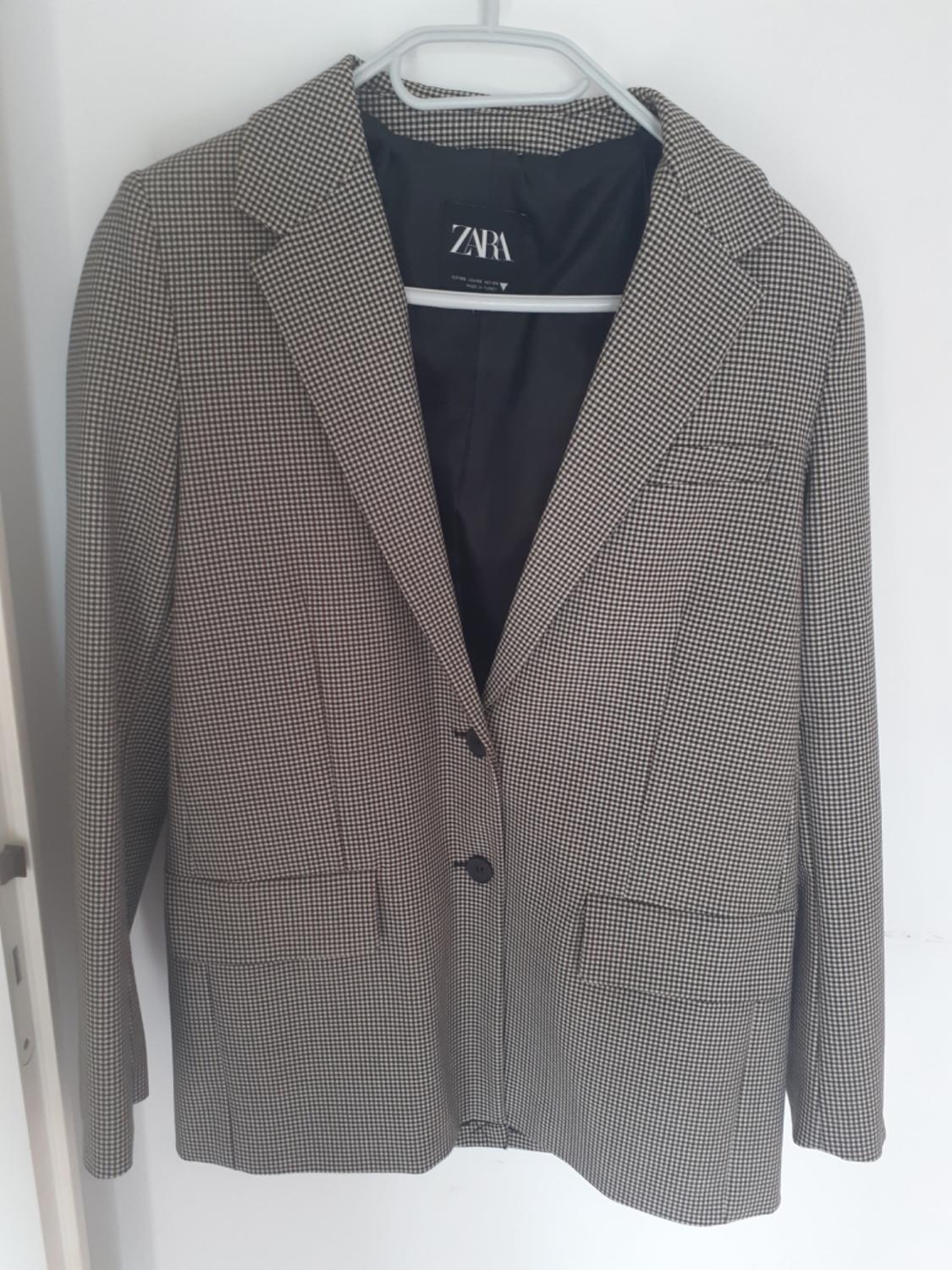 Checked blazer with two buttons hanging on a white hanger against a light background. Casual style. Black lining with the ZARA text.