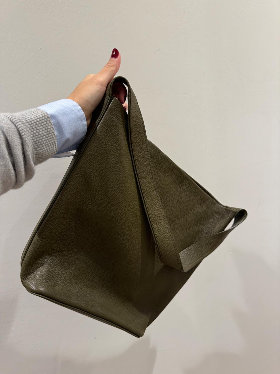 Hand holding a brown leather handbag with a minimalist design. Neutral background. Red painted nail and grey jumper with blue shirt.