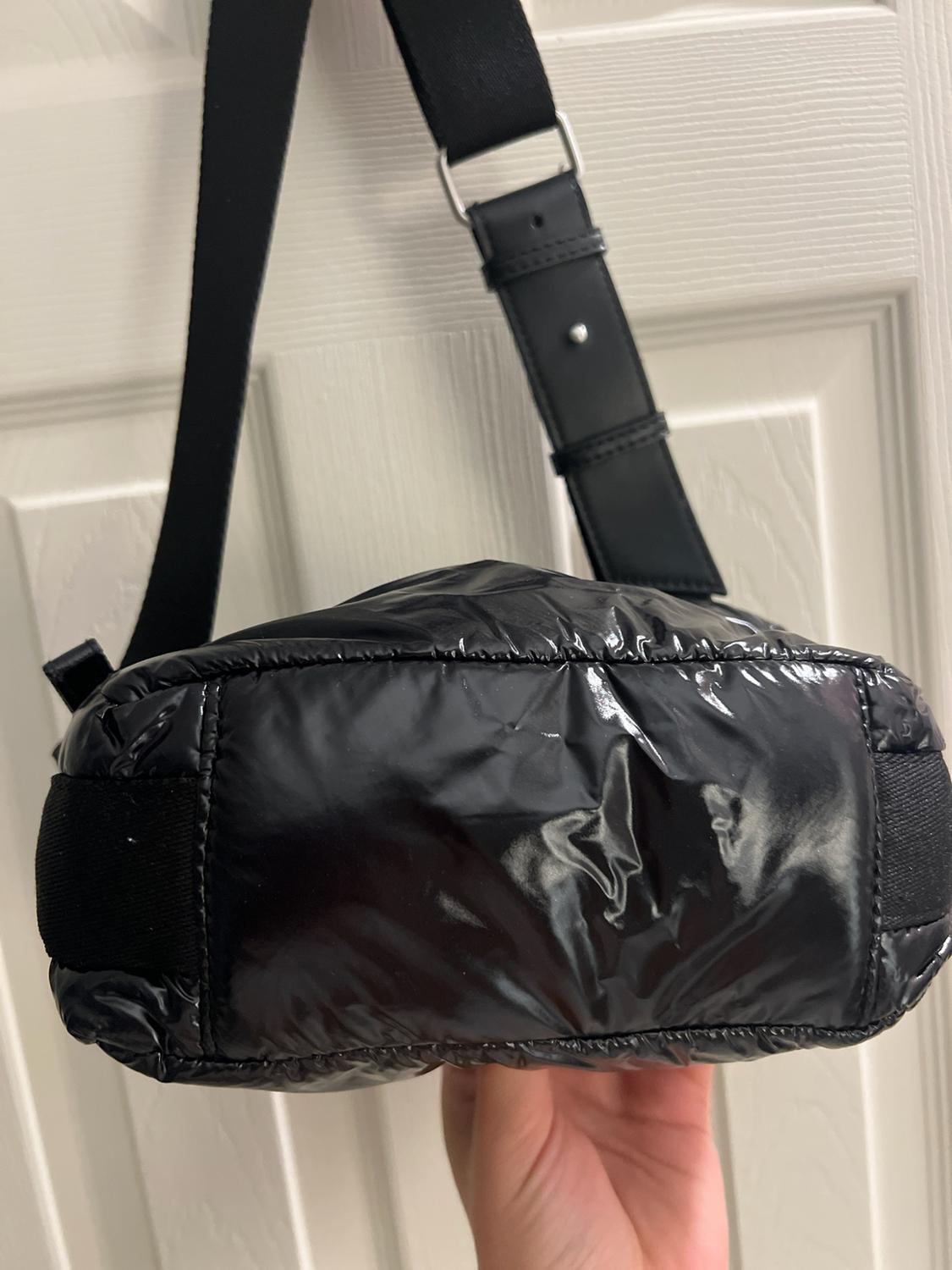 Hand holding a shiny black quilted bag by the base, with a wide black strap, in front of a white door.