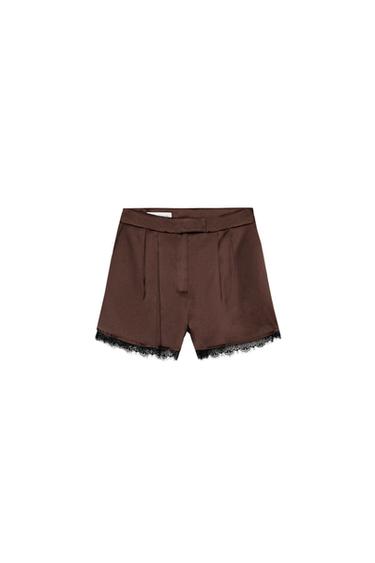 Front view of brown Bermuda shorts with front pleats, side pockets, and black lace trim. Front zip fastening.