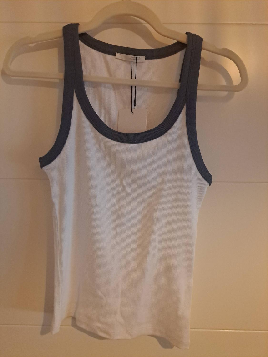 White ribbed vest top with dark grey trim, hanging on a white hanger against a wall of white panels.