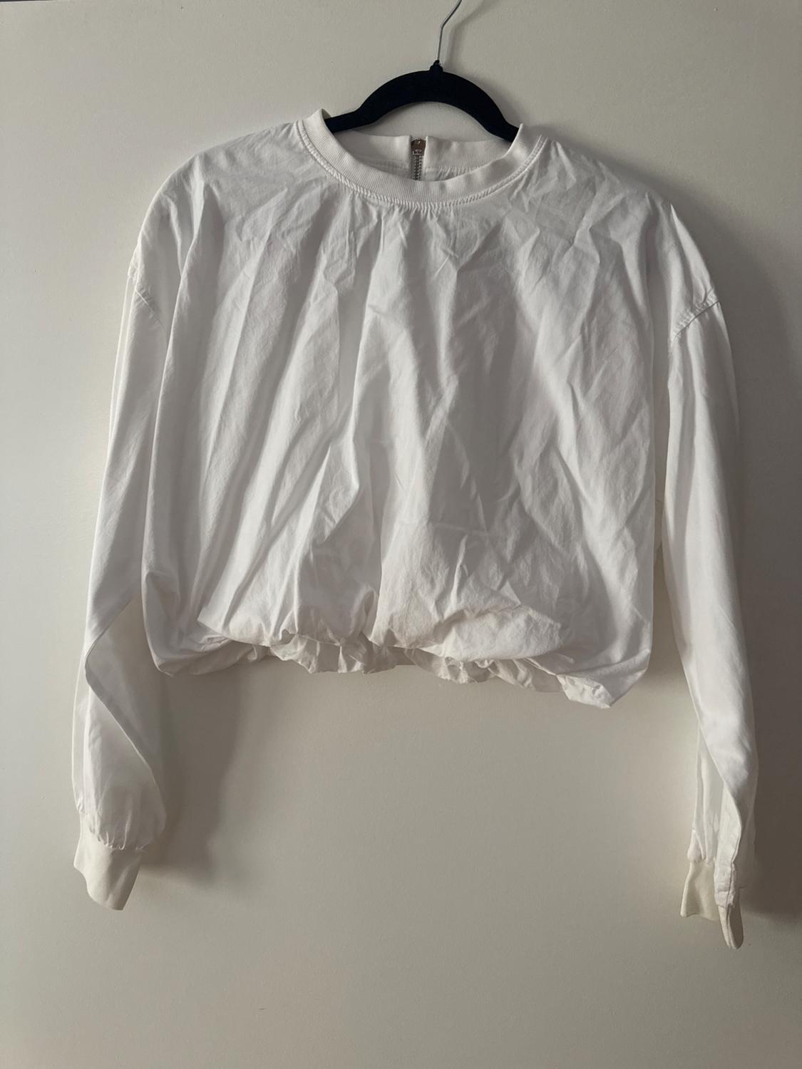 White long-sleeved top, round neck with zip and gathered hem, hanging on a black hanger against a white wall. Relaxed style.