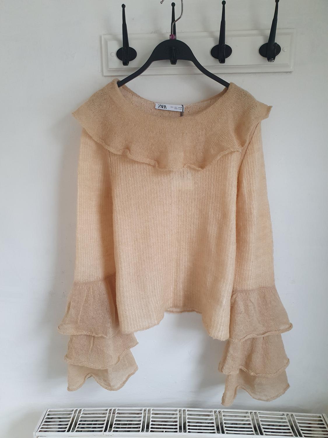 Beige jumper with frills on the sleeves and collar, hanging on a black hanger against a white background. Visible label: ZARA.