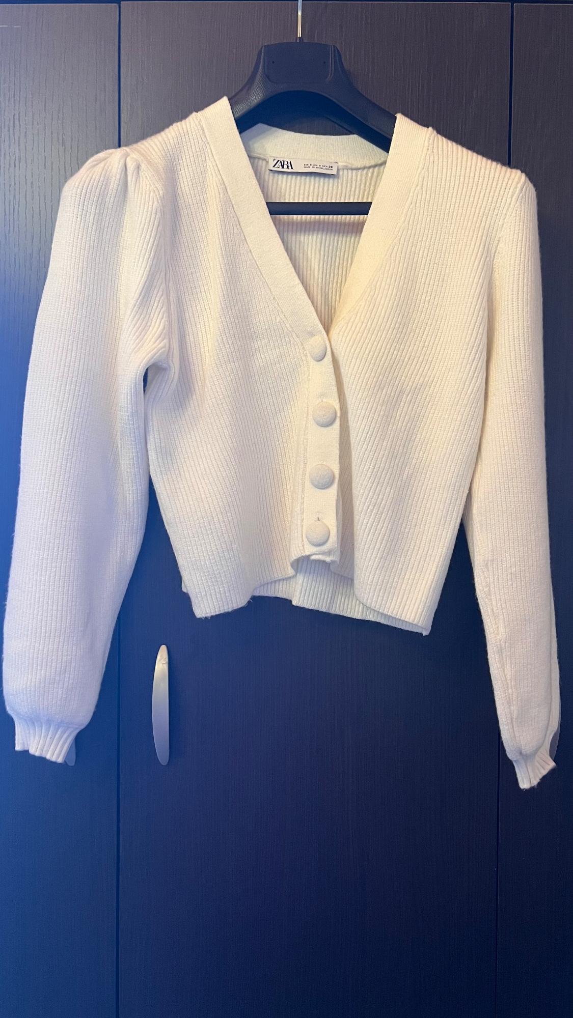 White knitted cardigan with large buttons, hanging on a black hanger in front of a dark wardrobe. Visible label: ZARA.