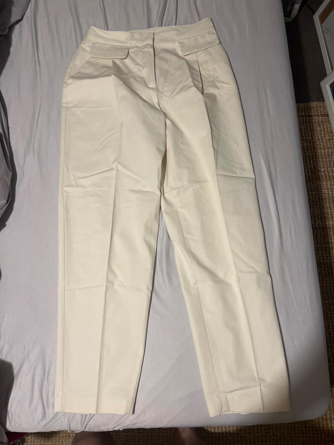 Cream-coloured trousers with pleats and waist tabs, laid out on a bed with grey sheets in a relaxed and casual setting.