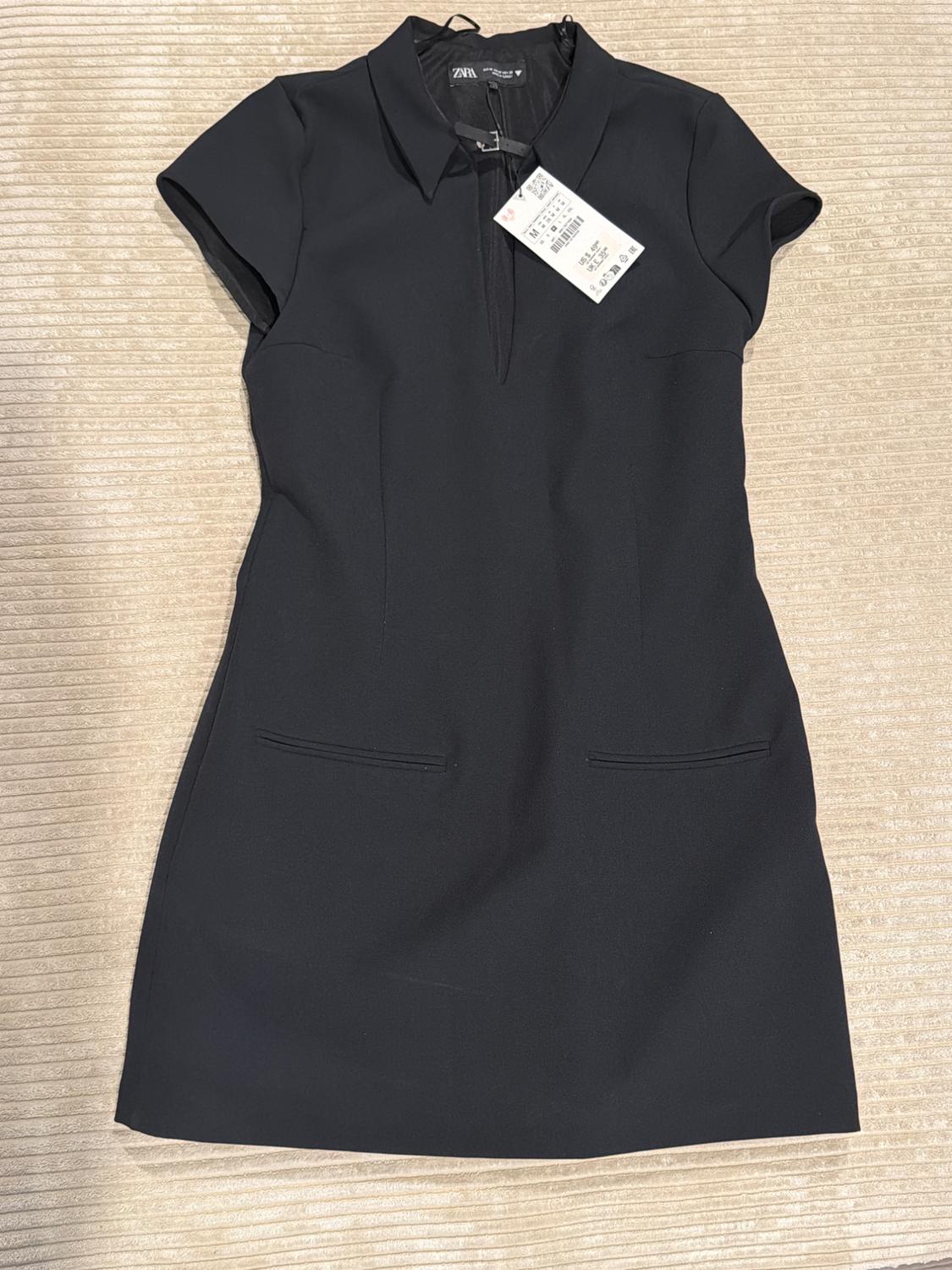 Short black dress with short sleeves, a collar and V-neckline, front pockets, and a ZARA label, laid out on ribbed beige fabric.