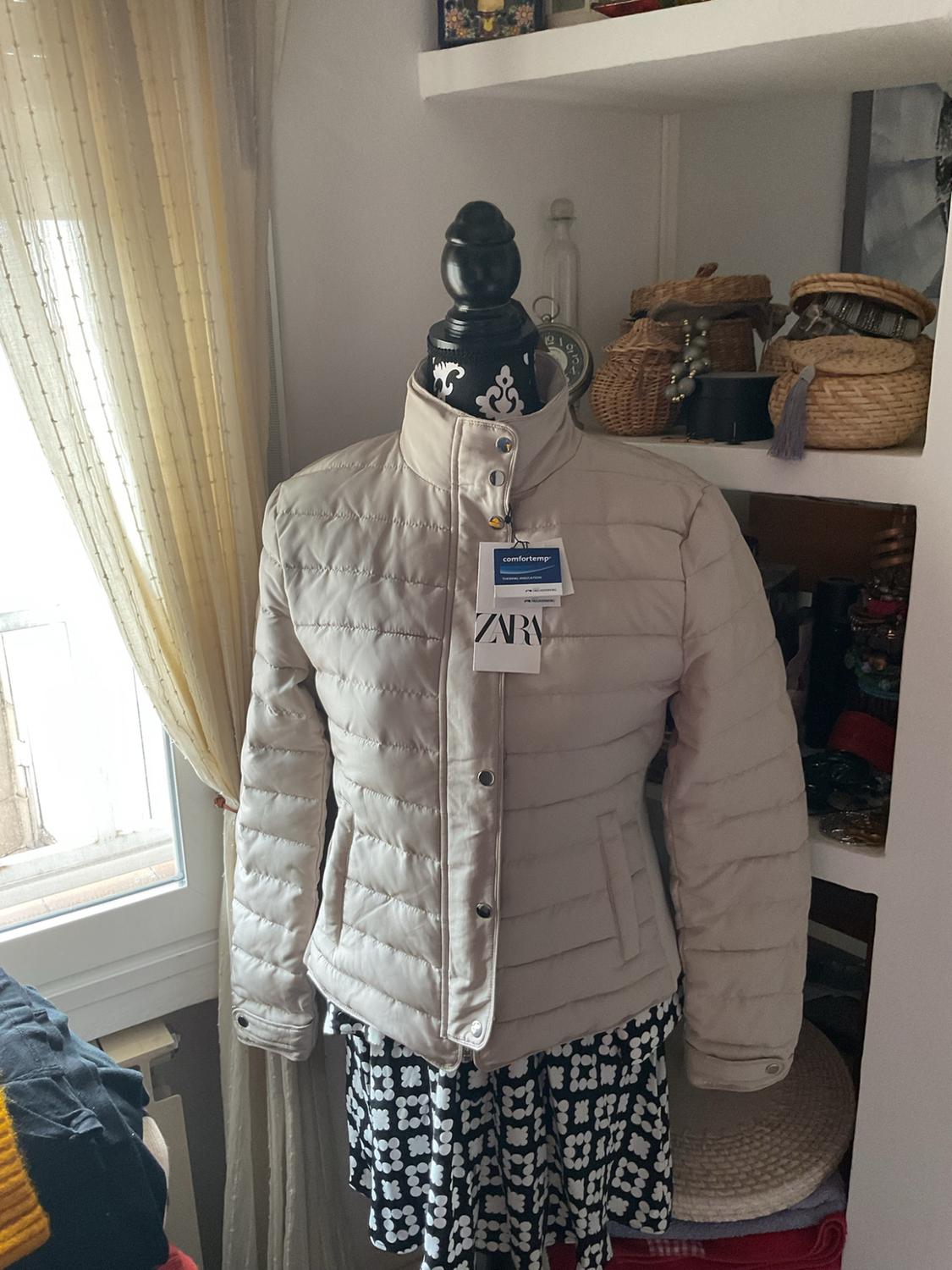 Mannequin wearing a beige quilted jacket, “comfortemp” and “ZARA” labels, and a black-and-white patterned skirt, in a room with shelves.