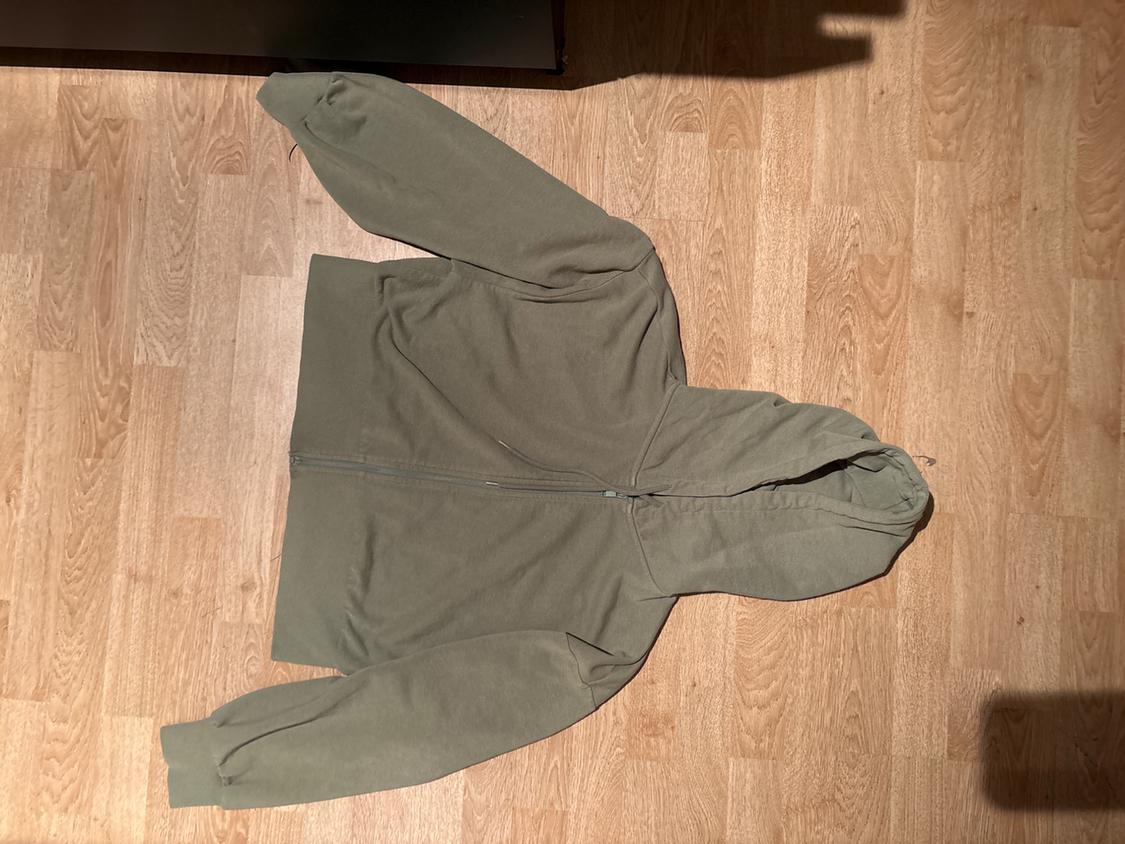 Olive green hoodie on light wooden flooring. Long sleeves and front zip fastening.
