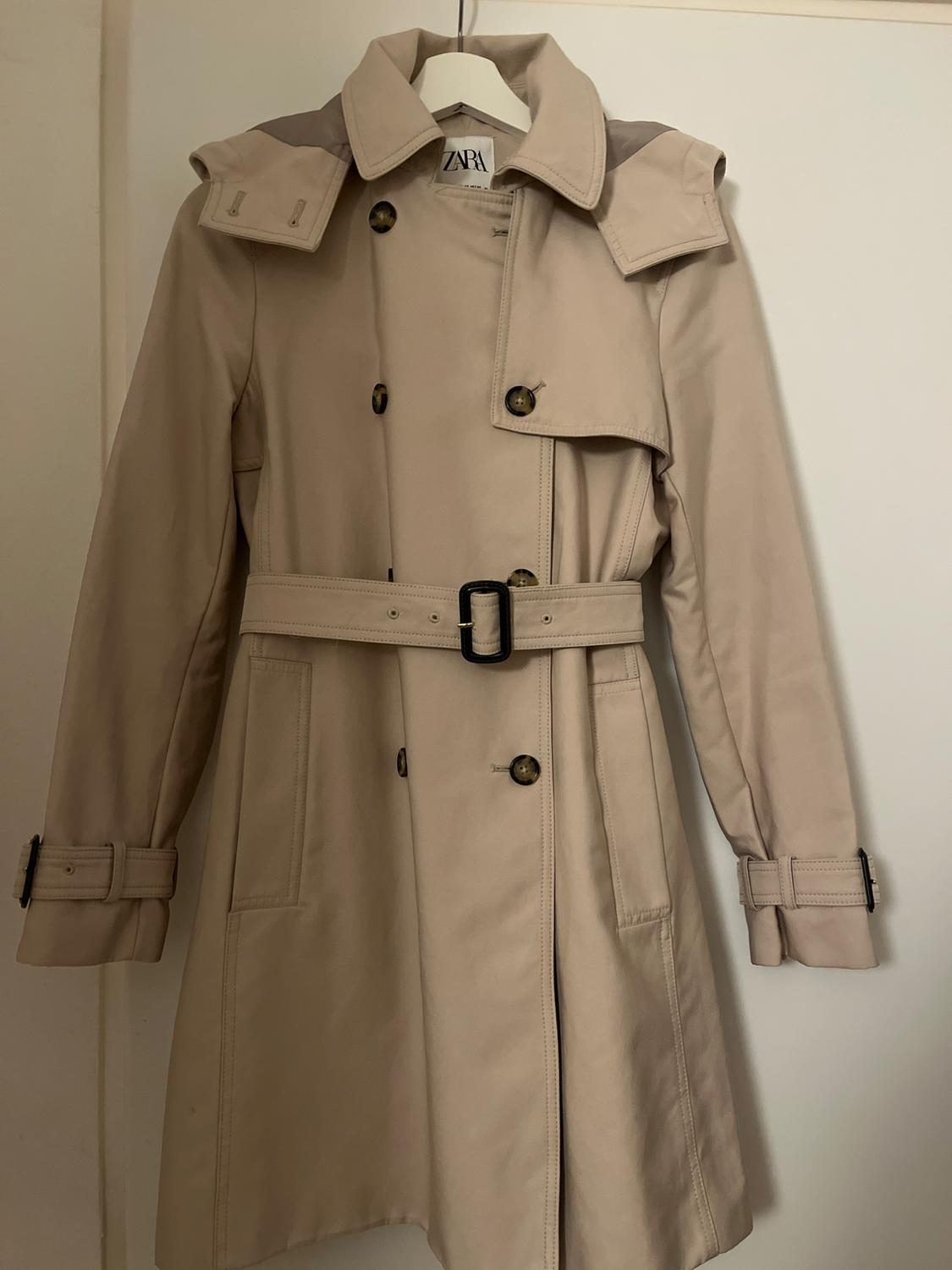 Beige trench coat hanging on a white hanger, with a belt and dark buttons. Visible label with the text "ZARA".