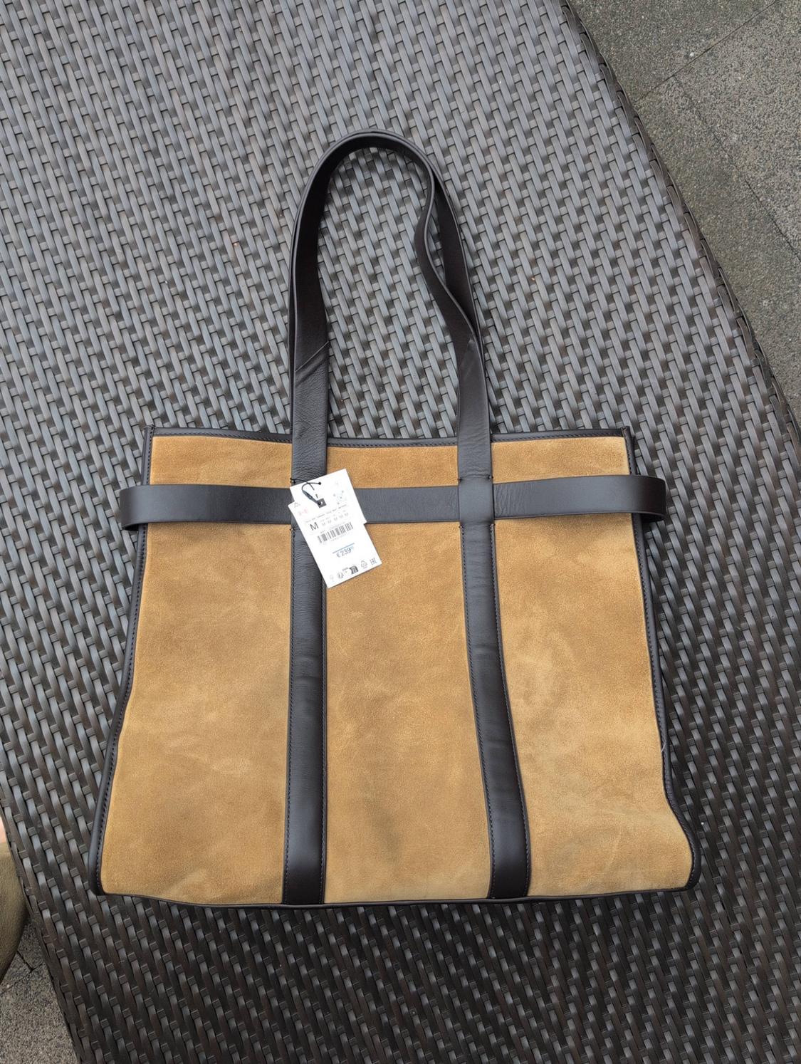 Tan-coloured suede tote bag with brown handles, resting on a woven wicker chair outdoors. Label with the text M £239.