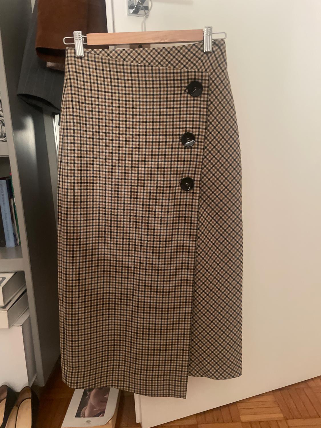 Brown and black checked skirt with large buttons on the side, hanging on a wooden hanger in a wardrobe.