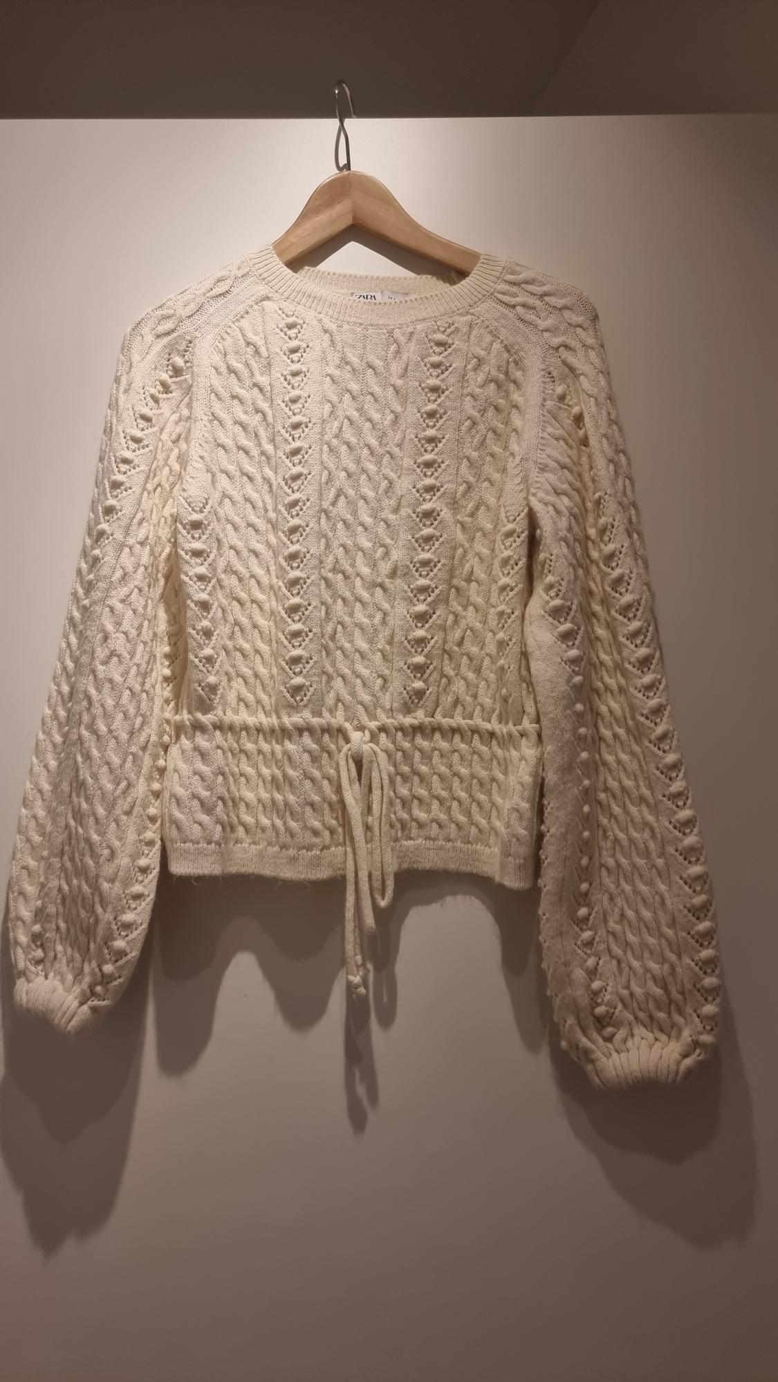 Cream knitted jumper hung on a wooden hanger, with cable knit design and adjustable drawstring waist.