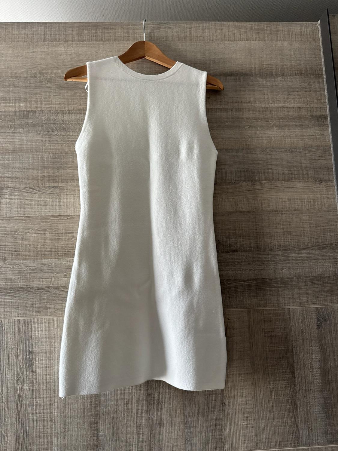 Short white sleeveless dress hanging on a wooden hanger against a wood panel background. Minimalist and casual style.