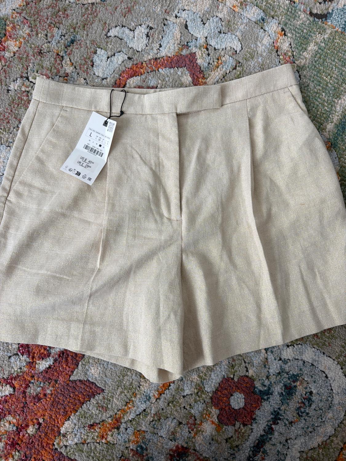 Beige dress shorts with pleats on a patterned rug. Tag with text: L US $45.90 UK £29.99.