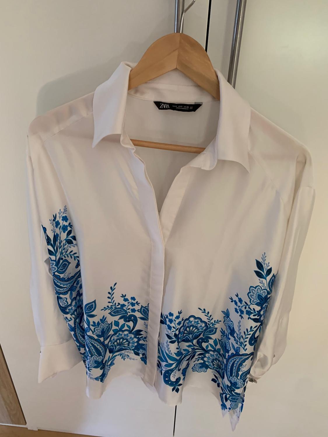 White long-sleeved shirt with a blue floral print, hanging on a wooden hanger. Inner label with the text ZARA.